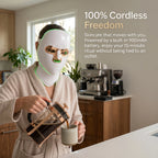 Woman pouring coffee in a modern kitchen while wearing a cordless, battery-powered LED facial mask, demonstrating a 100% hands-free morning skincare routine.