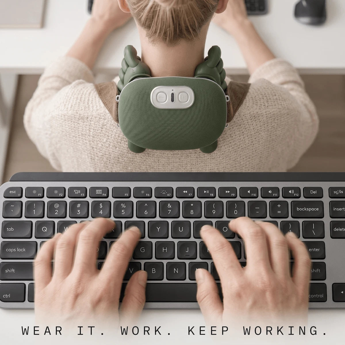 A professional typing on a laptop at a minimalist office desk while wearing a hands-free sage green neck massager, showing both hands completely free for work.